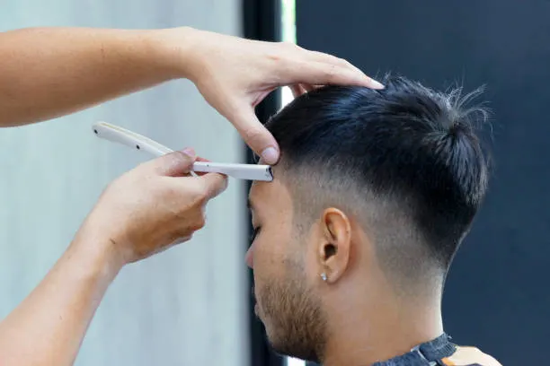 Barber make a haircut for customer at barbershop. Using razor blade. Small and retail business.
